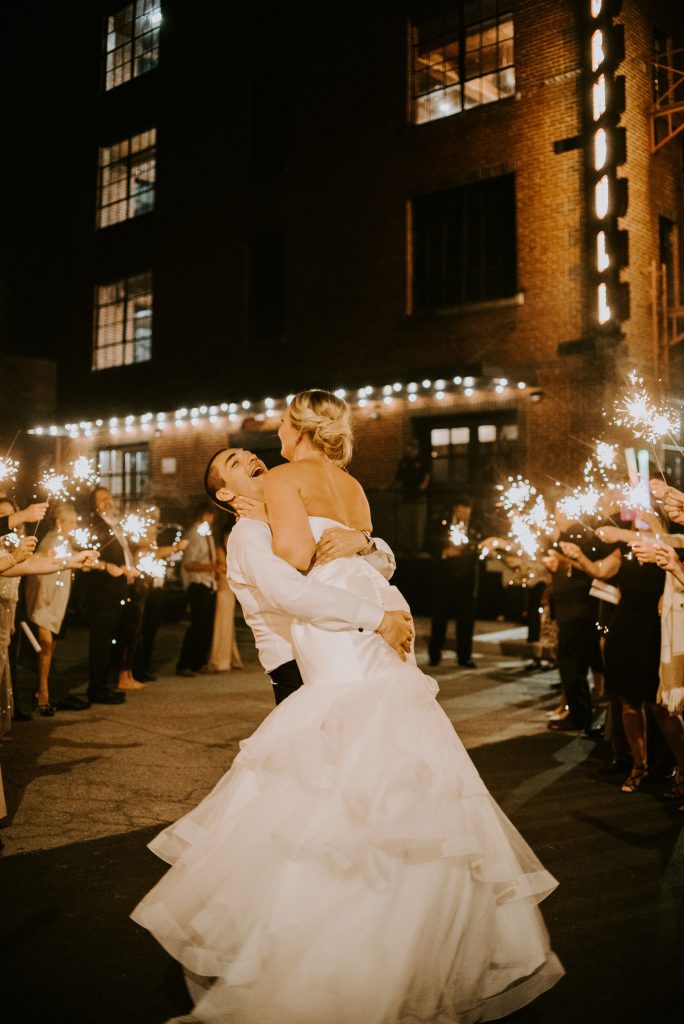 The groom lifts his bride in celebration as they leave their industrial chic wedding to head to their honeymoon. 